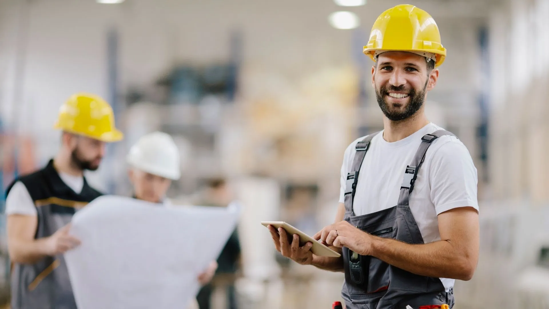 Elektrotechniker hält ein Tablet in der Hand und lächelt in die Kamera, währendessen 2 Kollegen im Hintergrund einen Plan betrachten