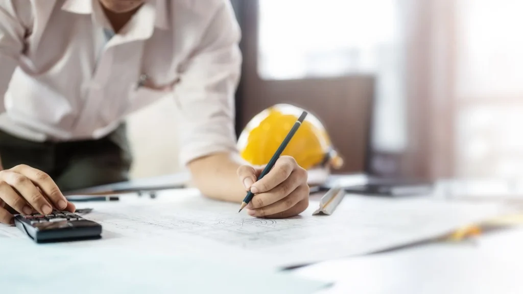 Eine Person ist über eienn Bauplan am Schreibtisch gebückt und tippt in etwas in den Taschenrechner ein.