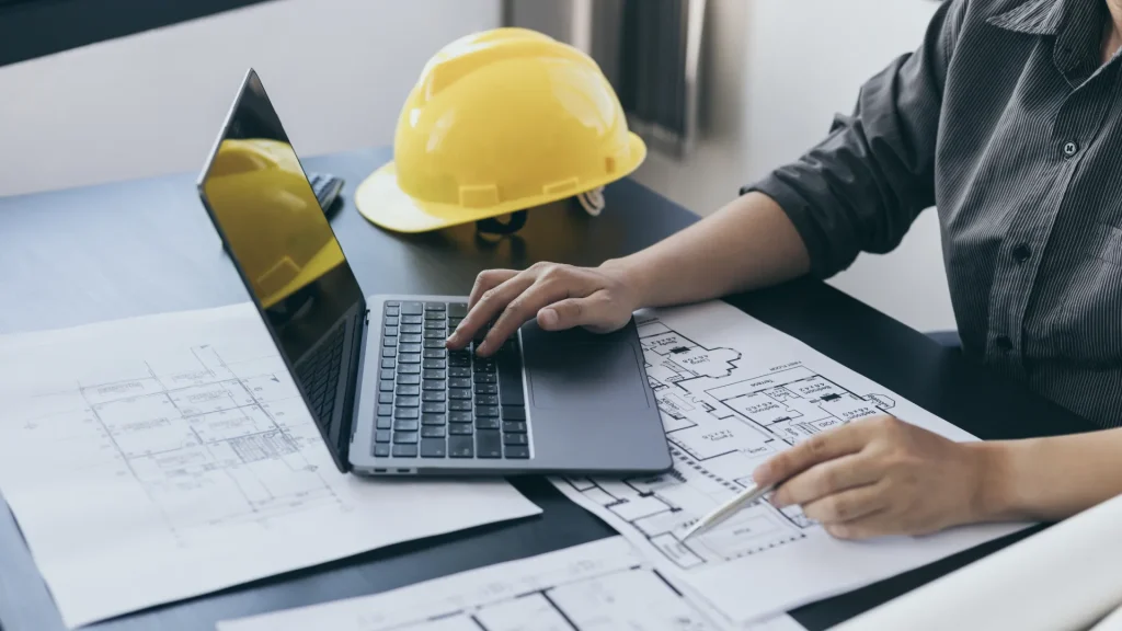 Sparolin-Elektrotechnik-Elektroinstallation - Eine Person sitzt am Schreibtisch, auf dem Baupläne, ein Laptop und ein gelber Helm ausgebreitet sind.