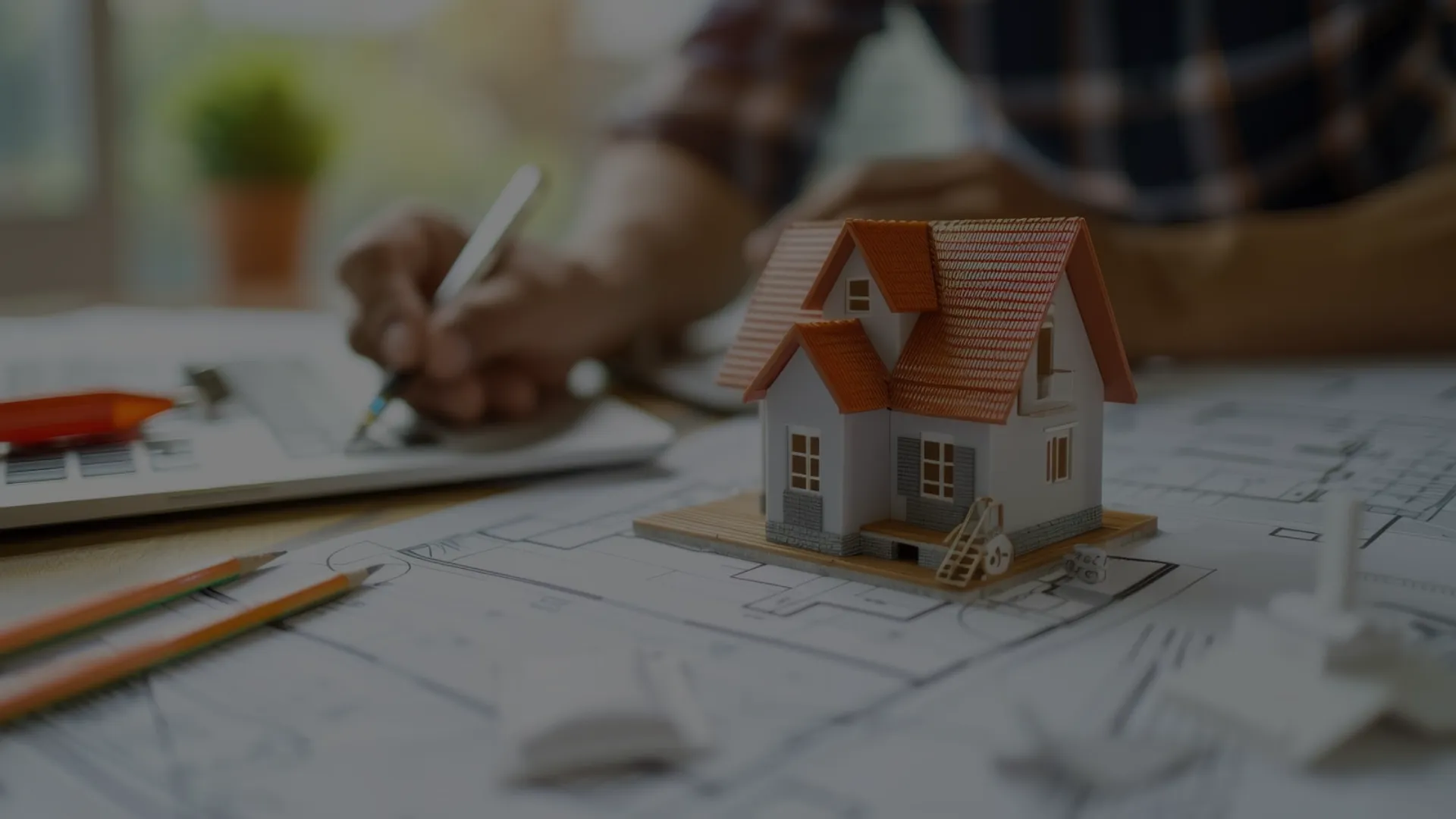 Ein Haus steht auf einem Bauplan. Im Hintergrund sieht man einen Mann, der auf eine Block schreibt