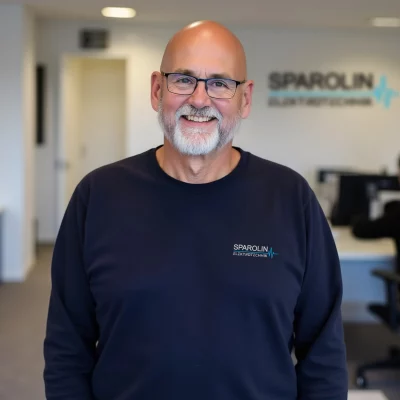 Der Geschäftsführer von Sparolin-Elektrotechnik Thorsten Sparolin steht lächelnd vor einer Wand mit dem Firmenlogo und blickt in die Kamera